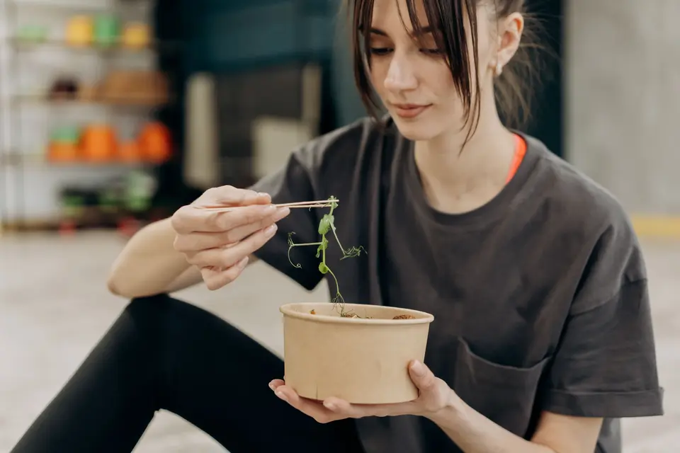 A woman sitting on the floor eating from a kraft paper bowl with chopsticks