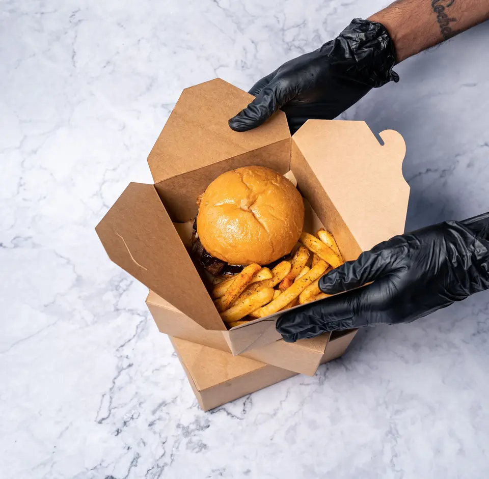 Gloved hands holding an open kraft takeout box with a burger and fries