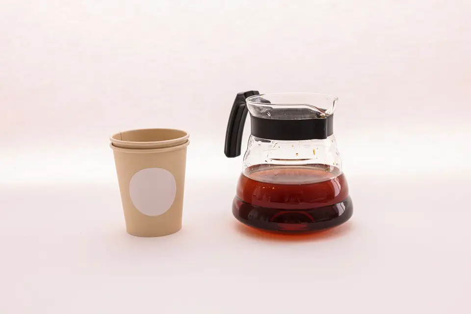 A kraft paper cup next to a glass carafe of coffee