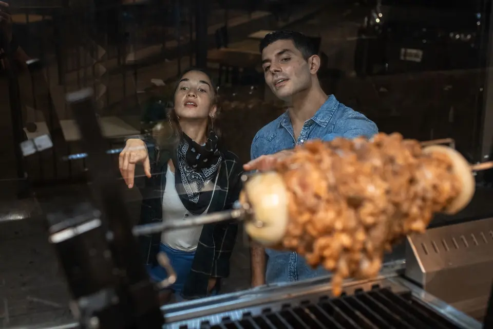 Two customers at a food truck window watching meat turn on a spit